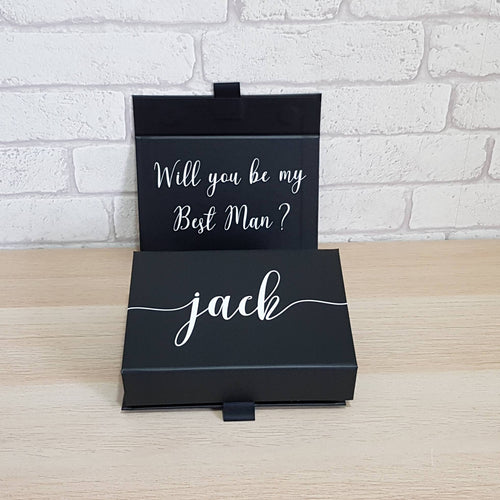 small black rectangular gift box with a proposal in white text on the inside of the lid. A name across the front in white. 